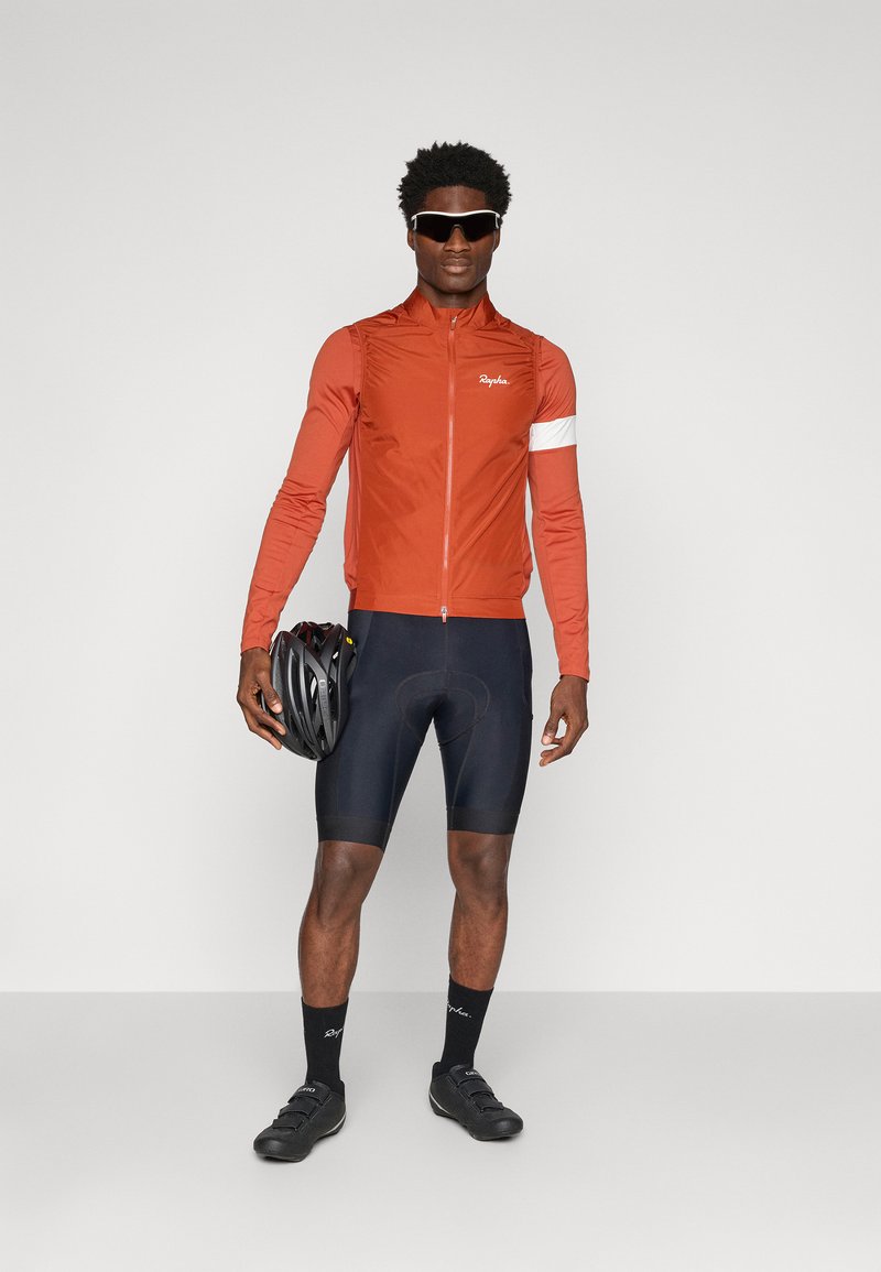 Männliches Radsport-Outfit mit einer taillierten, roten Windjacke mit weißen Akzenten, schwarzen gepolsterten Shorts und schwarzen Radschuhen. Hält einen Fahrradhelm.