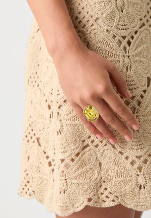 Bague en cristal jaune de forme octogonale avec un design facetté, portée sur une main au-dessus d'une robe en crochet beige texturée.