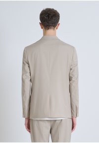 Veste de costume beige ajustée avec une fente unique au dos, des manches longues et des poignets boutonnés. Tissu lisse avec une texture minimale.