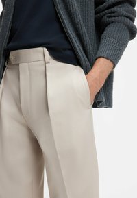 Beige pleated pants with a smooth texture, paired with a dark blue shirt and a textured gray cardigan featuring visible stitching.