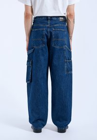 Lösa cargobyxor med vida ben i mörkblå denim, med flera stora fickor, synlig söm och en avslappnad passform. Bakvy som visar midjebandet med etikett.
