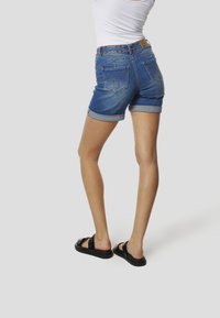 Közép kék farmer rövidnadrágok felhajtott szegéllyel, két hátsó zsebbel és barna bőr címkével a derékrésznél. Fekete szandállal párosítva.