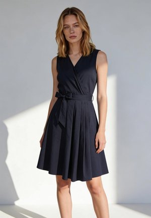 Frau trägt ärmelloses schwarzes Wickelkleid mit gebundenem Taillengürtel, steht vor schlichtem hellem Hintergrund mit Schatten an der Wand.