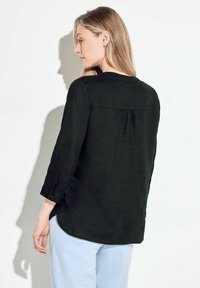 Blusa nera, strutturata, con scollo rotondo e maniche a tre quarti. Il retro presenta una leggera piega. Abbinata a pantaloni azzurri chiari.