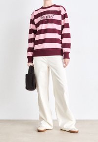 Sudadera con rayas burdeos y rosas con el logo "tommy jeans", combinada con pantalones anchos color crema y zapatos planos beige. Sosteniendo un bolso gris oscuro.