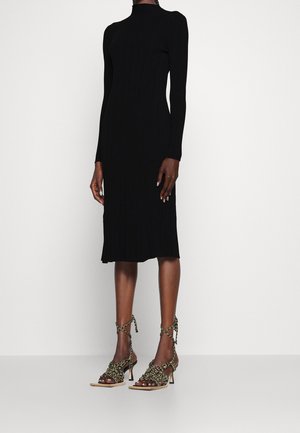 Frau trägt ein langärmeliges, knielanges schwarzes, geripptes Kleid mit hohem Kragen, kombiniert mit beigen und schwarzen geflochtenen Sandalen mit hohem Absatz.