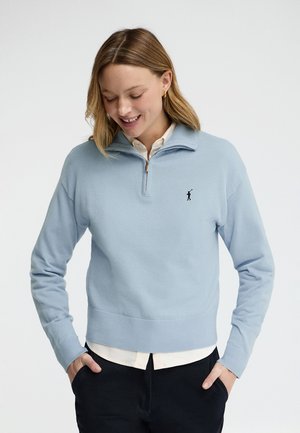 Jonge vrouw die een lichtblauwe trui met halve rits draagt over een wit shirt, met de handen in de zakken van haar zwarte broek, glimlachend en naar beneden kijkend.