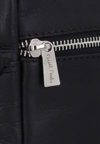 Reißverschluss einer schwarzen Ledertasche mit silberner Metallbeschlägen, versehen mit einem Anhänger mit der Aufschrift "Gusti Leder" und einer strukturierten Lederoberfläche.