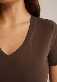 Hnědý tričko s výstřihem do V a krátkými rukávy, vyrobené z hladké látky. Výstřih má mělčí tvar písmene V, odhalující část klíční kosti.
