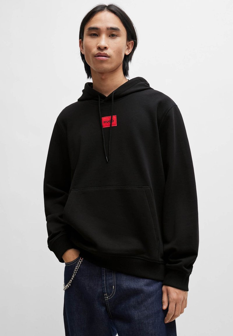 Svarte hettegenser med frontlomme og rødt logo-patch. Har snor til hetta og ribbede mansjetter. Båret over mørkeblå jeans.