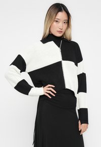 Sweater cropped às riscas preto e branco com fecho em zíper e textura canelada. Apresenta um colarinho e mangas longas, usado por cima de um vestido preto.