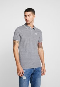 Grå polo skjorte med teksturert stoff, kontrasterende hvit krage og ermet trim, med en liten logo på brystet. Bært med slitt jeans.