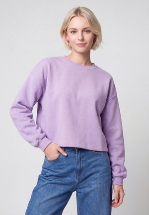Ung kvinde med kort blondt hår iført en lilla langærmet sweatshirt og blå jeans, stående med den ene hånd i lommen.
