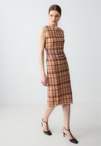 Frau, die in einem ärmellosen beigen und braunen karierten Midikleid mit gerafften Seiten und schwarzen T-Strap-Highheels vor einem einfarbigen grauen Hintergrund geht.