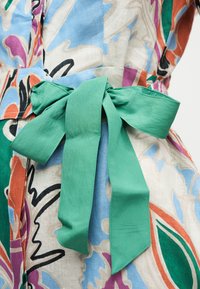 Ceinture en tissu vert nouée en noeud papillon sur une robe à motifs floraux multicolores avec des tons de bleu, orange, violet, vert et beige.