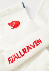 White socks with a ribbed design, featuring a red bear logo and "FJÄLLRÄVEN" in red text. Accented with blue and yellow stripes at the top.