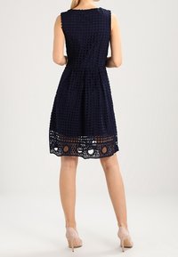 Námořnická modrá krajková šaty s přiléhavým živůtkem, rozšířenou sukní, vlnitým lemem a kruhovými výřezy na spodní části. Bezrukávový design pro klasický vzhled.