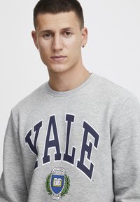 Sudadera gris con "YALE" en grandes letras azules marinas y un escudo con un libro y hojas de laurel. Cuello redondo clásico y textura suave.