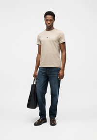 Μπεζ βαμβακερό t-shirt με το λογότυπο "Tommy Hilfiger", μπλε τζιν τζιν, και καφέ δερμάτινα παπούτσια, κρατώντας μια μαύρη τσάντα tote. Απλός σχεδιασμός.