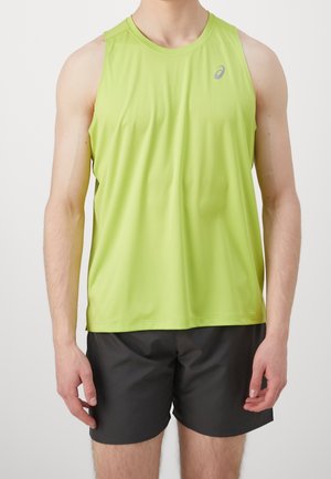 Man draagt een limegroen mouwloos sporttopje en zwarte short, staat met armen ontspannen langs het lichaam tegen een effen achtergrond.