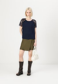 Mornarska bluza z čipkastimi rokavi, olivno zelena krila, rjavi škornji do kolen in bela torbica. Glatke teksture, preproste oblike in kontrastne barve.