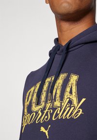 Marineblå hættetrøje med et stort slidt gult "PUMA sports club" grafisk motiv og matchende snor med metalender.