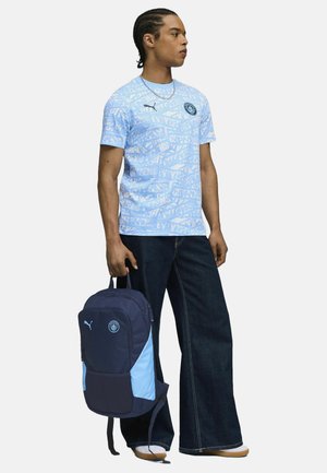Maglietta grafica azzurro chiaro con motivo di testo ripetuto, abbinata a jeans larghi scuri; tiene uno zaino blu navy e azzurro chiaro.