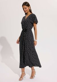 Kobieta ubrana w czarną sukienkę midi w białe groszki, z rozkloszowanymi rękawami, taliowaną talią, srebrne szpilki, perłowa bransoletka i duże kolczyki.