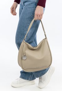Beige leather shoulder bag with a slouchy shape, tiny metal tag, and strap details; worn with light blue high-waisted jeans and white sneakers.