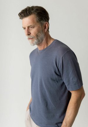 Uomo di mezza età con capelli grigi e barba, che indossa una camicia blu a maniche corte, che guarda pensieroso verso il basso su uno sfondo chiaro e semplice.