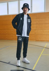 Tuta nera con logo NBA Nets e branding Nike, abbinata a sneaker bianche; all'interno, con pareti in legno sullo sfondo.