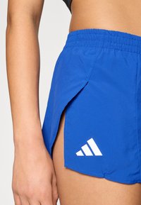 Pantalones cortos deportivos azules con cintura elástica, aberturas laterales y detalle de logotipo en forma de triángulo blanco. Fabricados con tela ligera y suave.