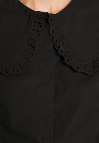 Blouse noire avec un col rond orné de dentelle délicate, fermeture à boutons au niveau du cou et une texture de tissu lisse.