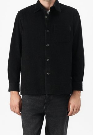 Homme portant une chemise en velours côtelé noire à boutons avec poche poitrine et un jean gris foncé, montré du cou aux hanches.