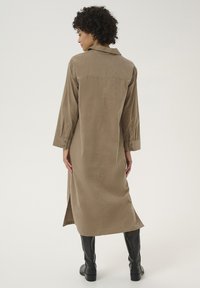 Abito camicia lungo color khaki realizzato in tessuto morbido, con colletto, polsini bottonati, fianchi spacchettati e silhouette dritta. Indossato con stivali neri al ginocchio.