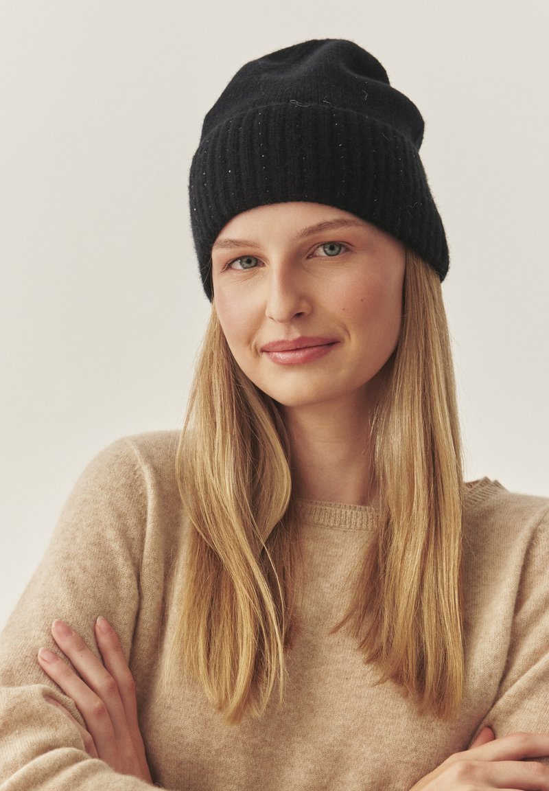 Mustast soonikudumist müts soonilise tekstuuriga. Pehme, soe kangas. Kandmine helebeeži kampsuniga. Modellil on pikad juuksed ja lõdvestunud asend.