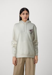 Crème hoodie gemaakt van zachte stof, met een voorzak en een klein grafisch logo in bordeauxrood, gedragen over beige broeken.