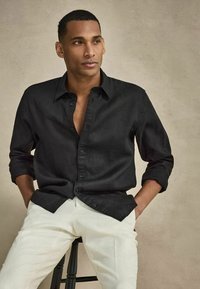 Homme aux cheveux courts portant une chemise noire boutonnée et un pantalon blanc, assis sur un tabouret noir devant un fond beige.