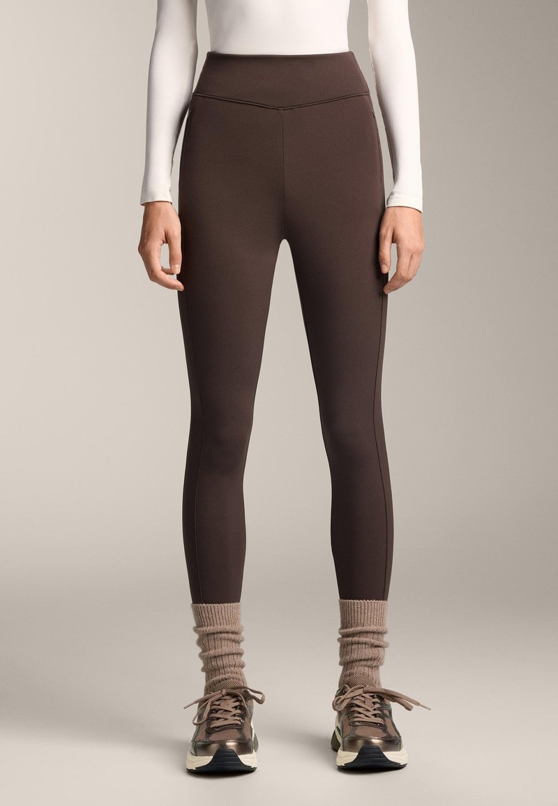 Bordeaux hoge taille leggings gemaakt van gladde, rekbare stof met contrasterende stiksels. Gecombineerd met grijze gewatteerde sokken en sportieve schoenen.