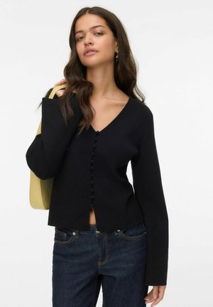 Maglione nero con scollo a V, maniche allungate e una fila di bottoni rotondi sul davanti. Indossato con jeans scuri e una borsa di colore chiaro.
