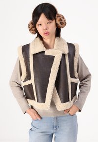 Brun faux lædervest med beige pelsfor, der har en åben front, struktureret design og bred krave. Modellen har en grå sweater og jeans på.