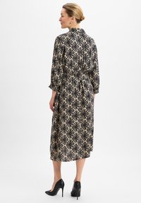 Schwarzes und beiges Kleid mit floralem Muster, langen Ärmeln, taillierter Taille, gerader Silhouette und knielangem Saum. Kombiniert mit schwarzen High Heels.