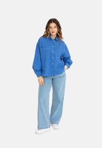 Niebieska koszula zapinana na guziki z dwoma kieszeniami na piersi, luźny krój i długie rękawy, w połączeniu z jasnoniebieskimi szerokimi spodniami dżinsowymi i białymi sneakersami.