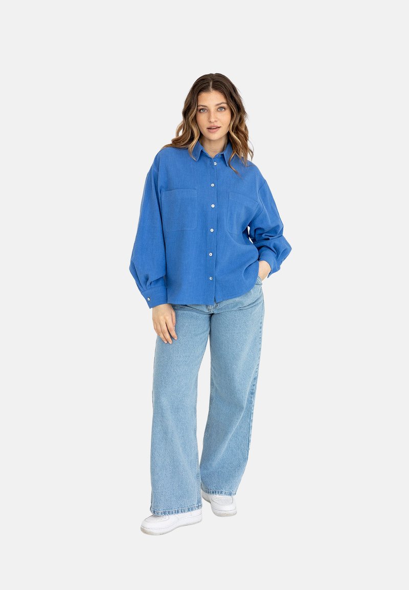 Niebieska koszula zapinana na guziki z dwoma kieszeniami na piersi, luźny krój i długie rękawy, w połączeniu z jasnoniebieskimi szerokimi spodniami dżinsowymi i białymi sneakersami.