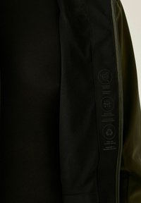 Primer plano del interior de una chaqueta negra que muestra el forro de malla, cremallera e íconos que representan características de impermeabilidad, transpirabilidad y materiales reciclados.