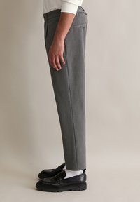 Grijze op maat gemaakte broeken met een fijn ruitpatroon, slim fit en zijzakken. G gedragen met zwarte instappers en witte sokken.