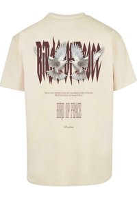 Creme bomulds T-shirt med et grafisk motiv af to duer med udstrakte vinger, tekst "FUGL AF FRED" i stiliseret bordeaux typografi.