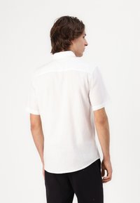 Camisa blanca de manga corta, hecha de tela ligera; presenta un cuello con solapa y un dobladillo ligeramente curvado en la parte posterior.
