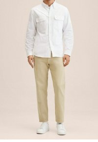 Valkoinen pitkähihaisella napilla varustettu paita, jossa on kaksi rintataskua, yhdistettynä beigeihin housuihin ja valkoisiin tennareihin, neutraalia taustaa vasten.