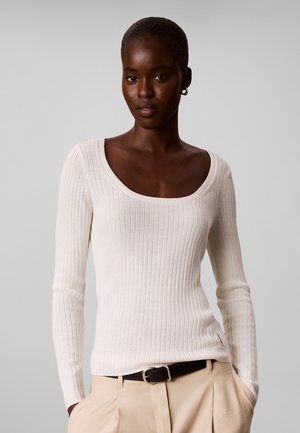 Frau mit kurzen Haaren trägt ein weißes, geripptes Langarmshirt und beige Hose mit schwarzem Gürtel, steht vor einem schlichten grauen Hintergrund.